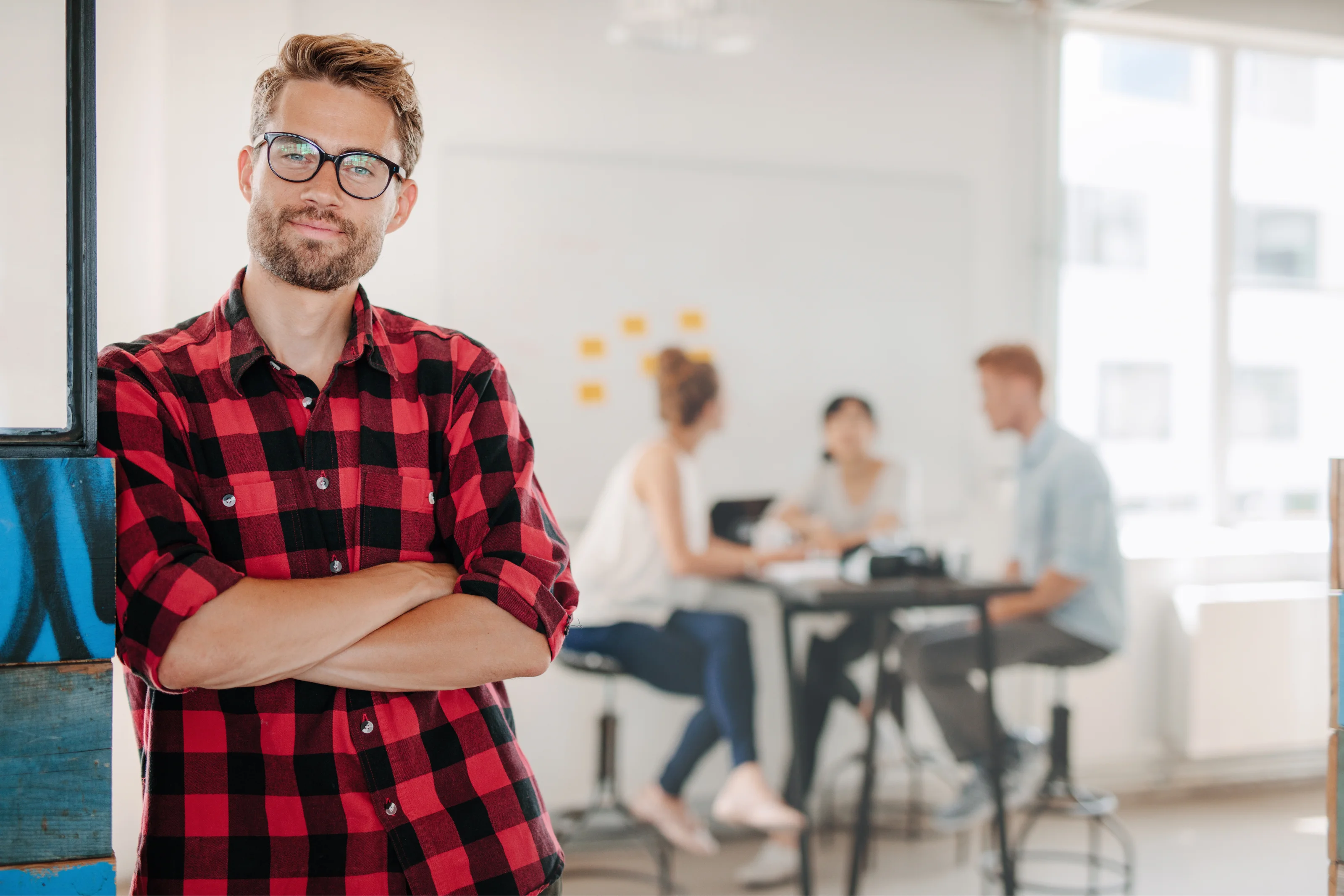 Uomo con camicia a quadri rossa appoggiato al muro, con un team che lavora sullo sfondo.