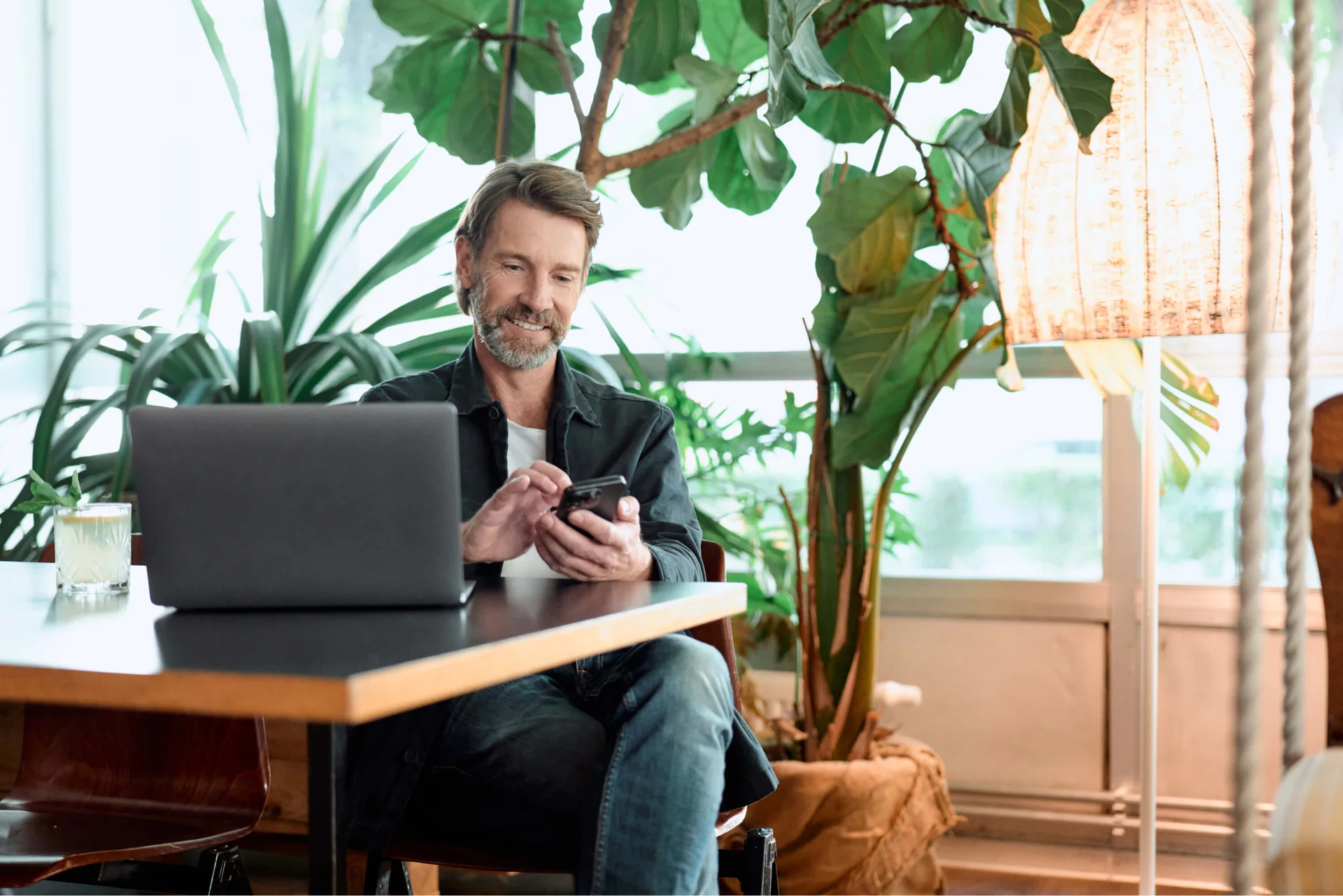 Uomo sorridente usa lo smartphone accanto al laptop in uno spazio pieno di piante