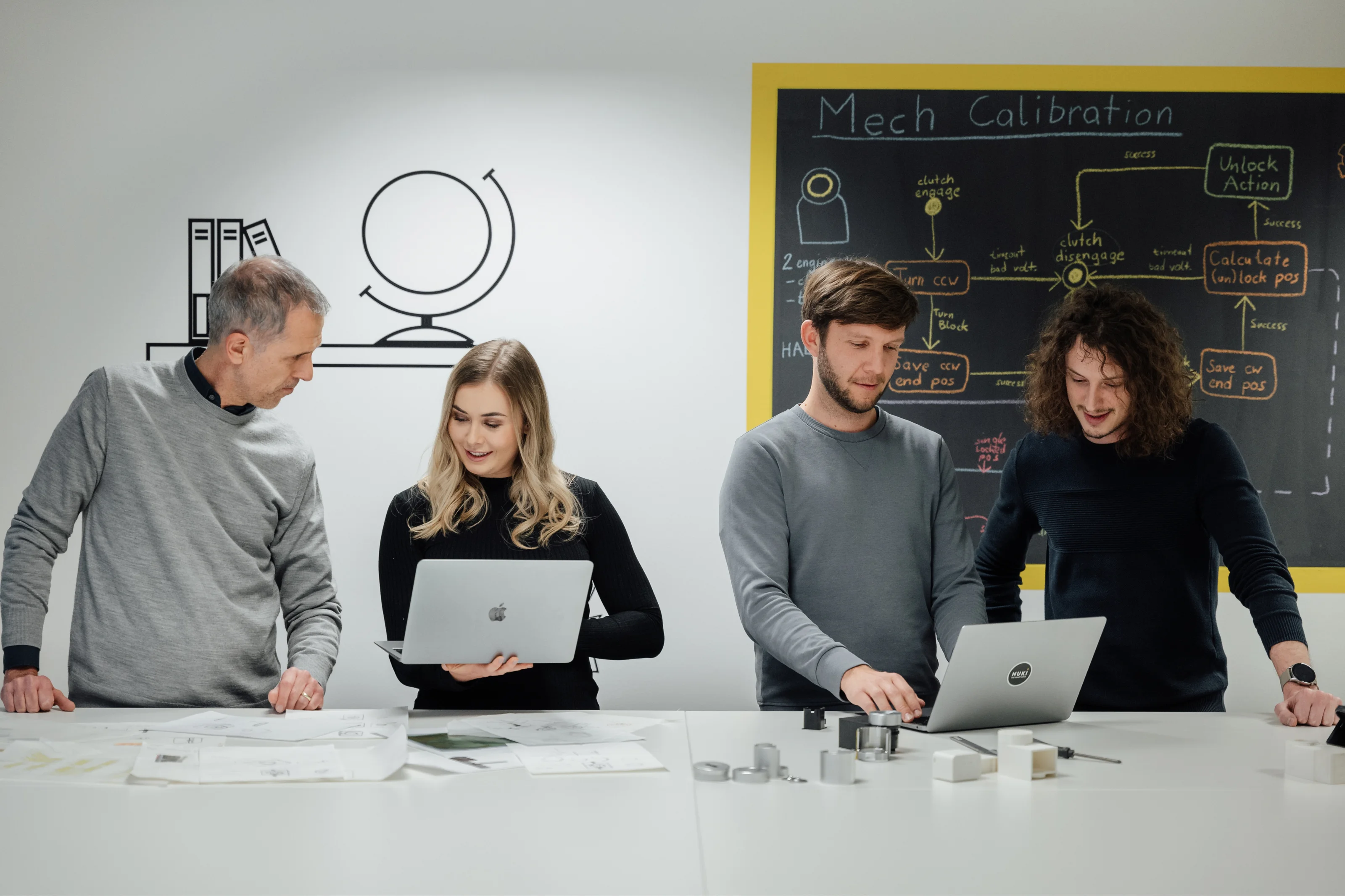 Leden van het Nuki-team werken samen voor een krijtbord tijdens een ontwikkelsessie met laptops en schetsen.