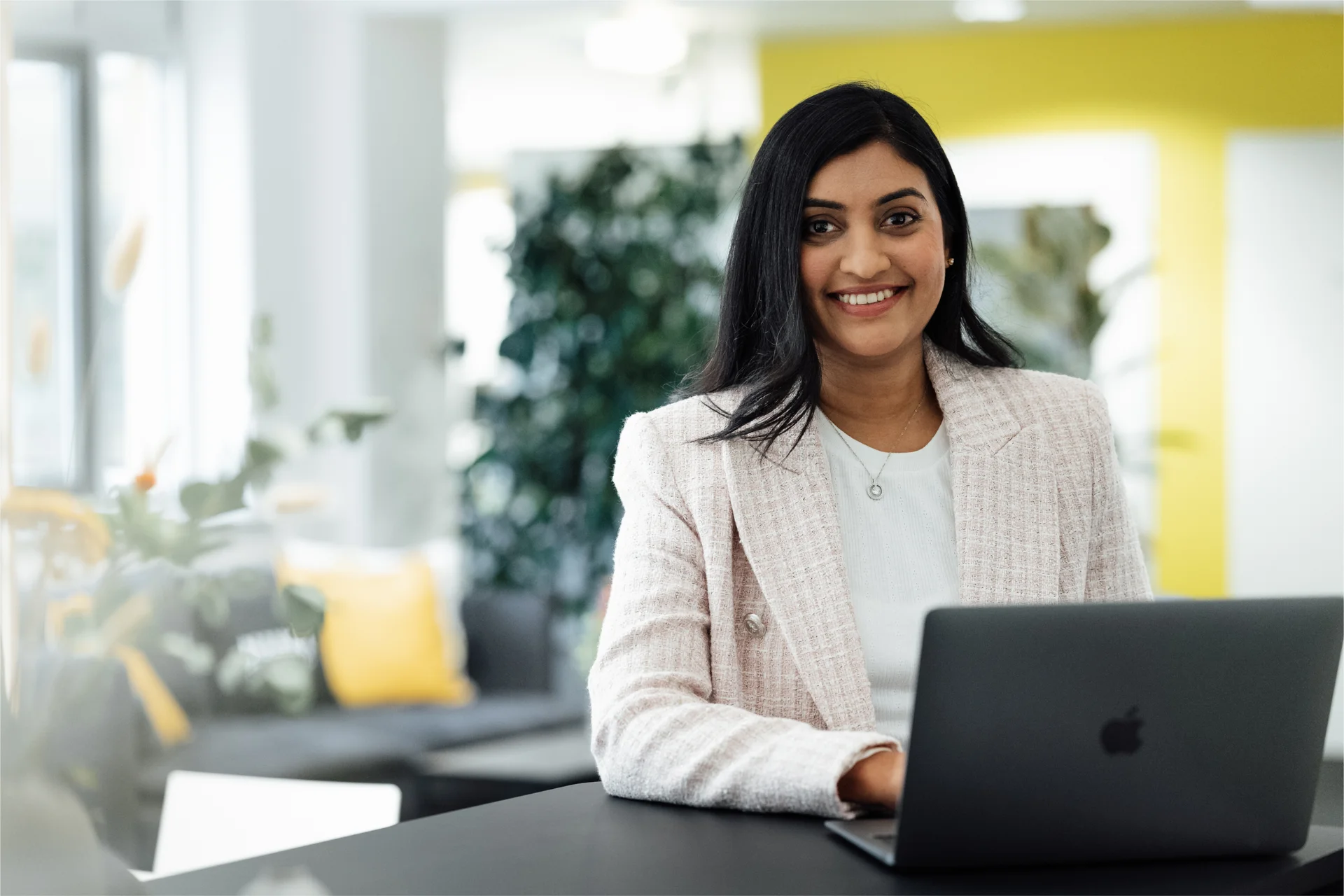 Glimlachend Nuki-teamlid werkt in een lichte kantoorruimte aan een donkere laptop, gekleed in blazer.