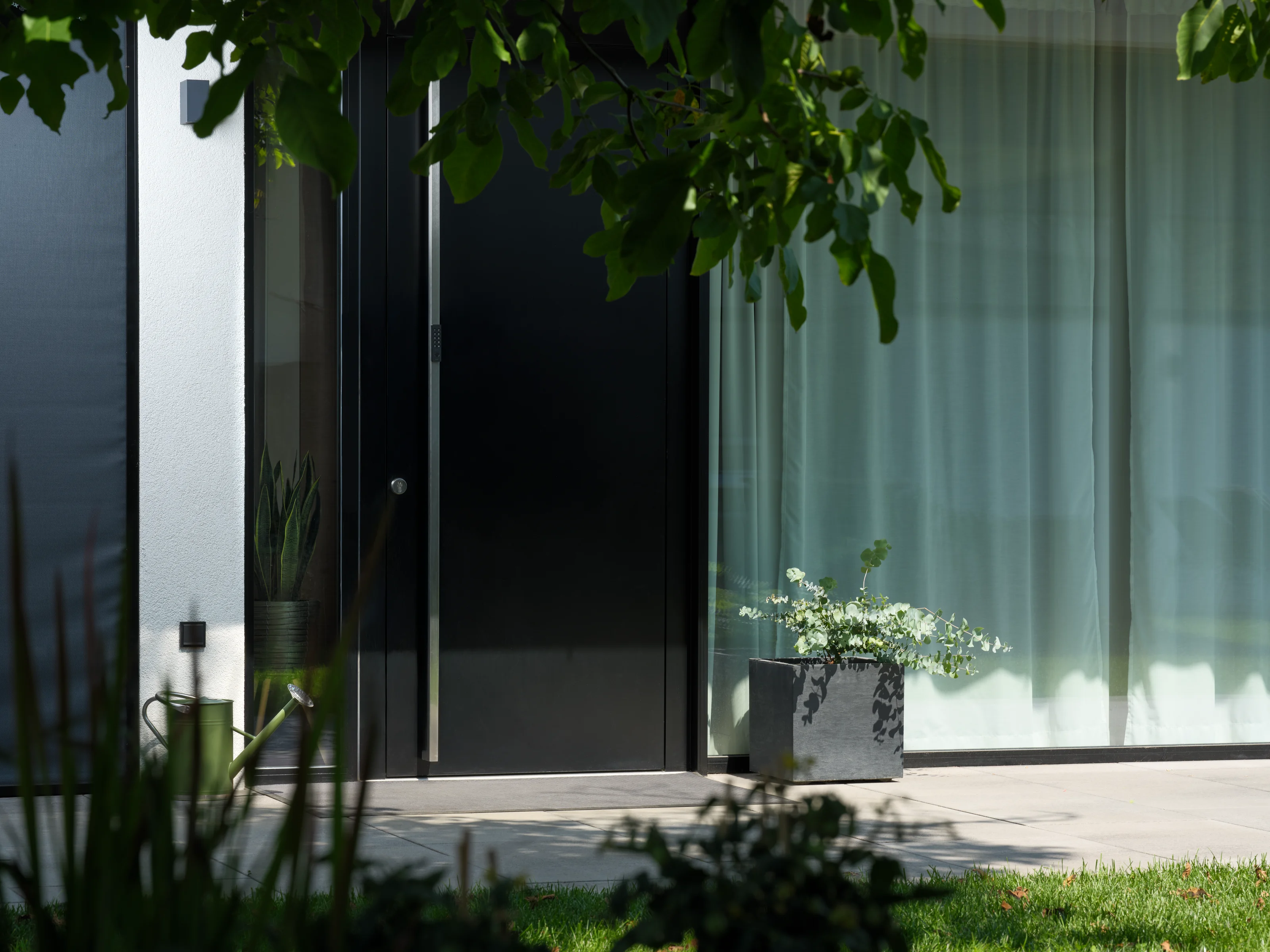 Moderna porta d’ingresso con ampie vetrate, circondata da piante e da un vaso con piante sulla terrazza.